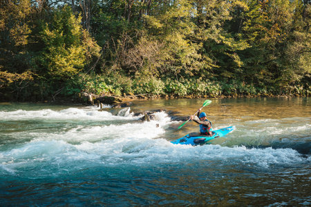 Young teenager cruising down whitewater rapids in a blue kayak, beautiful river nature on a sunny summer day, handheld shot.の写真素材