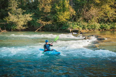 Young teenager cruising down whitewater rapids in a blue kayak, beautiful river nature on a sunny summer day, handheld shot.の写真素材
