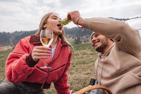 Smiling young modern couple having a trip in nature, enjoying wine and grapes while sitting relaxed next to their new exclusive electric carの写真素材