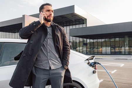 Businessman browsing on a smartphone while waiting to charge his electric car at a charging station in an airportの写真素材
