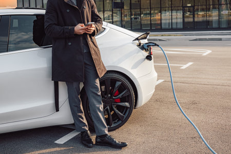 Businessman browsing on a smartphone while waiting to charge his electric car at a charging station in an airportの写真素材
