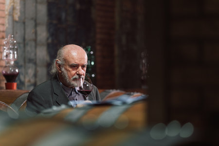 Senior graybeard Caucasian man, a wine connoisseur, holding a glass, analyzing the red wine through sight, smell, and tasteの写真素材