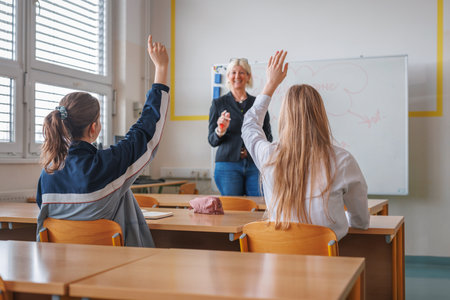 Girl student writes down notes and raises her hand while a female teacher lecturing in the class in front of the blackboard. Concept of classroom environment.の写真素材
