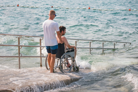 Man with disability at beach goes to swimm on a wheelchair with assistance on an accessible ramp.の写真素材