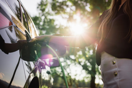 Female hand putting a cable charger in an electric car illuminated by the sun rays breaking through the branches of the leafy tree, close upの写真素材