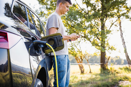 Young man charging electric car parked in the nature area and adjusting an EV charging app on smartphoneの写真素材