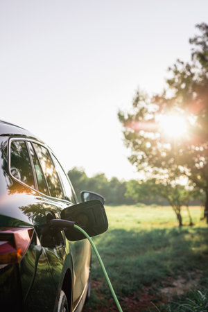Black electric car with the power cable supply plugged in, in the blurred background visible green area and trees. Eco friendly and renewable energy concept.の写真素材