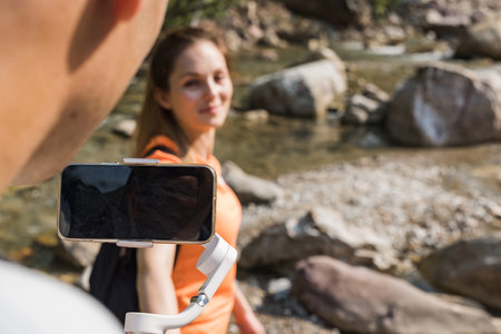 Handsome young couple standing in nature near the mountain river, making selfie vlog videos using a...