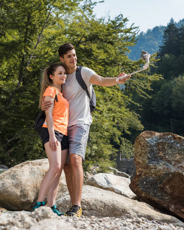 Handsome young couple standing in nature near the mountain river, making selfie vlog videos using a smartphone and selfie stickの写真素材