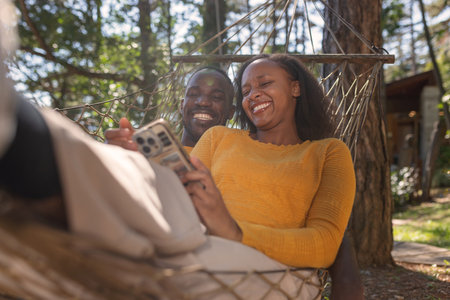 Cheerful African American couple relaxing in a hammock, watching funny videos on a smartphone, enjoying a glamping trip in Europe.の写真素材