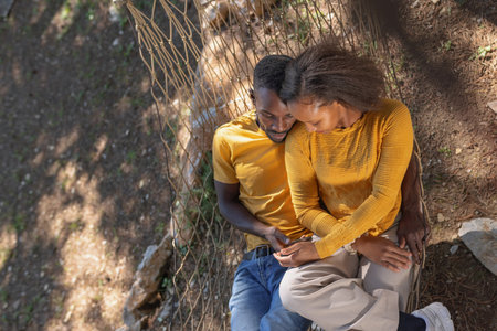 African American couple relaxing on a hammock, scrolling on smartphones, and enjoying nature outdoors in a glamping site in Europe.の写真素材