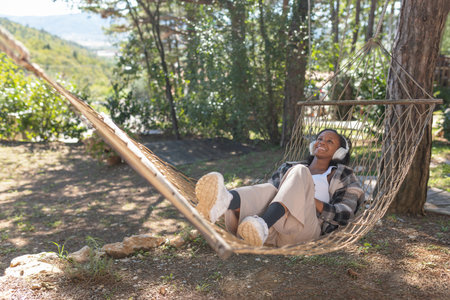 African American woman relaxing in nature on a hammock, choosing music on a smartphone and listening to favorite songs through headphones.の写真素材