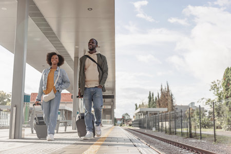 Young African American couple going on vacation, arriving at the railway station platform holding...