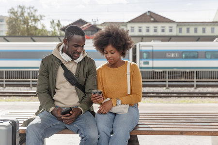 Smiling African American couple having fun using a smartphone while waiting for the train at the...
