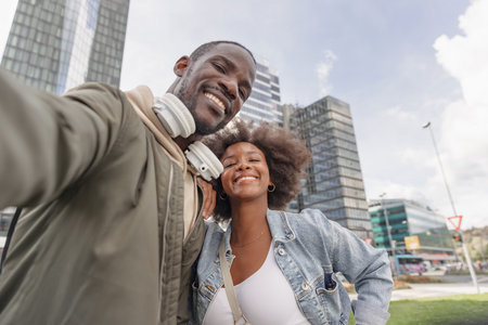 Cheerful African American couple excited about arrival at their travel destination, taking a selfie video in front of a modern hotel skyscraper.の写真素材