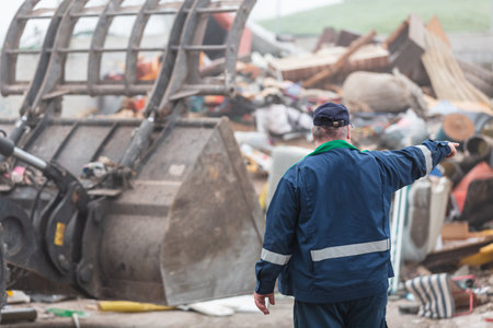 Landfill worker directing skid steer loader on the garbage heap, rear view. Waste disposal, consolidation, and transfer concept.の写真素材