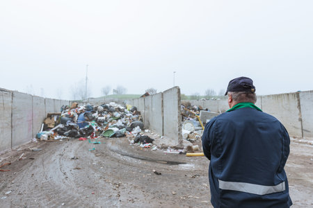 Recycling center worker, in dark blue work clothes, looking at an unsorted garbage heap just arrived in the materials recovery facility, rear view.の写真素材