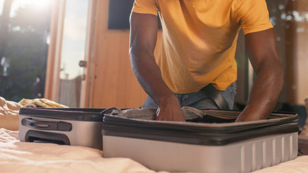 Smiling African American man packing his suitcase in a bedroom, preparing for travel, a long-awaited...