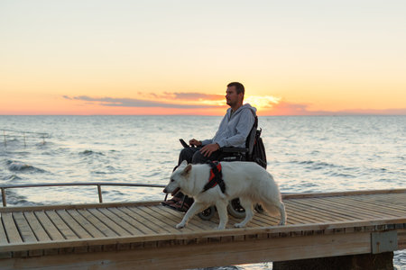 Man with disability moves along a wooden pier at sunset, accompanied by a white dog. The calm ocean...
