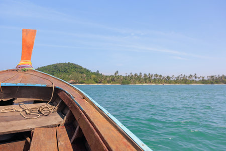 Bow of a wooden longtail boat cruising on beautiful, clear, turquoise water under a bright blue sky, with a green hilly island in the distance.の写真素材