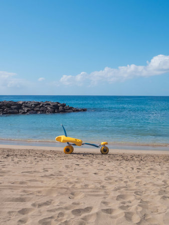 A beach wheelchair sits on the sandy shore, offering support for individuals with mobility challenges. The clear blue ocean and boats in the distance enhance the accessible atmosphere.の写真素材