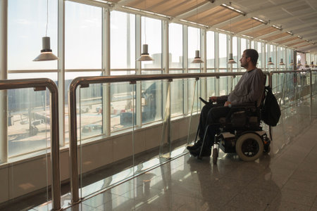 Person with disability using wheelchair observes the busy airport terminal from a vantage point. Bright sunlight streams in through expansive windows, highlighting the environment.の写真素材