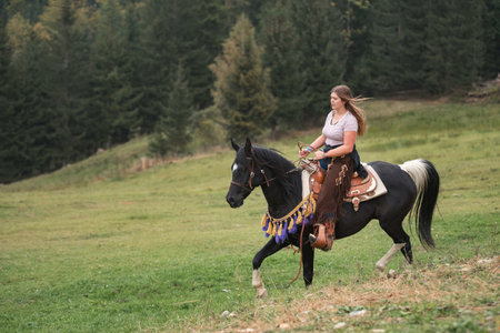 Confident young woman, a cowgirl with spirit of freedom and adventure riding a majestic black horse, through breathtaking mountain terrainの写真素材