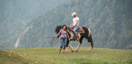 A man rides a black and white horse on a grassy hill, while a woman walks beside them holding the reins. Mountains are visible in the background.の写真素材