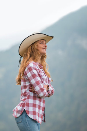A blonde woman wears a hat and a plaid shirt while standing near a mountain. She smiles and looks up with her arms crossed, enjoying the scenery.の写真素材