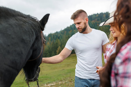 Cheerful young couple visiting a Western-style riding school, connecting with an instructor and a friendly horse as they feed it by handの写真素材