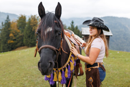 A young woman in western wear stands with her dark horse in a grassy pasture. Mountains and trees...