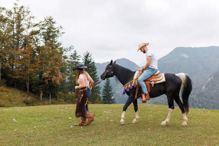 Cowgirl instructor, holding the reins, watching and helping a young rider mount his horse at the ranch riding schoolの写真素材