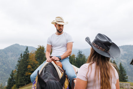 Authentic western riding school experience, cowboy learning horseback skills from a female instructor in a scenic mountain settingの写真素材