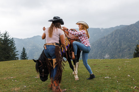 Two young women, dressed as cowgirls, prepare for a horseback ride on a rural hillside in the Austrian Alps.の写真素材