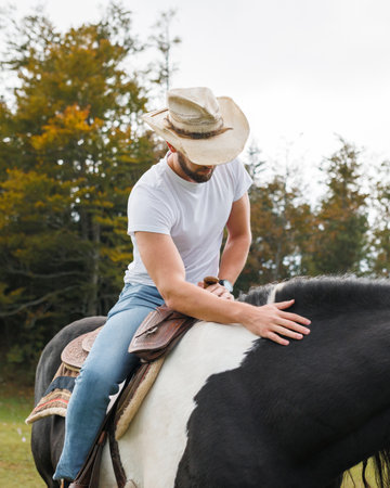 A man wearing a hat, t-shirt, and jeans is sitting on a horse outdoors. He is petting the black and white horse gently on its neck.の写真素材