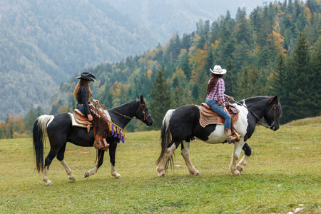 Two women wearing cowboy hats and western attire are riding black and white horses across a grassy field.の写真素材