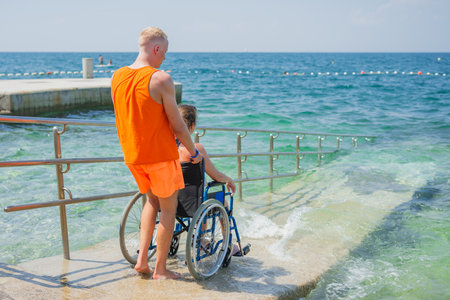 Woman with disability, entering the sea water with the help of a lifeguard assistant across an accessible wheelchair beach ramp, rear view.の写真素材