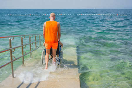 Lifeguard helping a woman with disability enter the water across the accessible wheelchair ramp on a public beach, allowing her to enjoy the sea.の写真素材