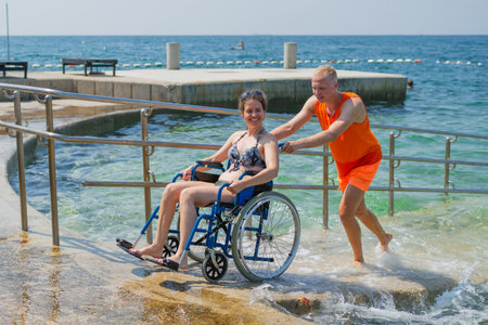 Relaxed woman with disability, exiting the sea water with the help of an assistant, across an...