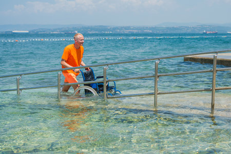 Smiling lifeguard rolling an empty aquatic wheelchair along an accessible beach ramp at a summer tourist destination with a beautiful seascape in the background.の写真素材