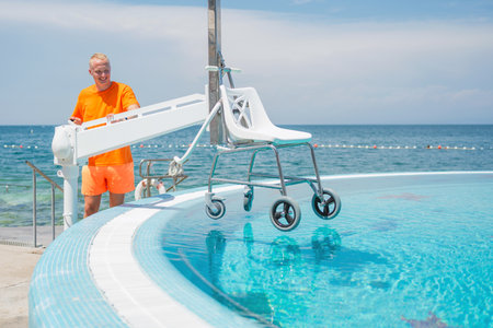 Man handling a pool chair lift, lifting and lowering from and into the water to provide access to the swimming pool for people with a physical disability.の写真素材