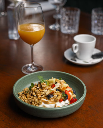 A bowl of yogurt topped with granola, fruits, and nuts alongside a glass of juice on a wooden table...