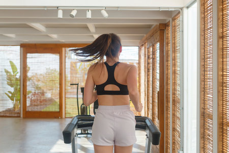 A woman runs on a treadmill in a well-lit exercise area with large windows and bamboo shades....