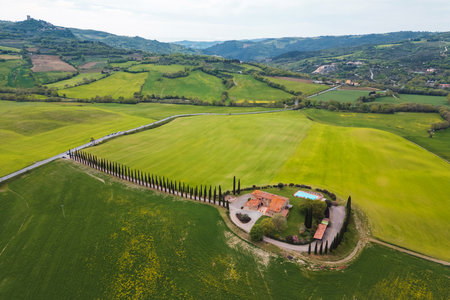 A charming villa surrounded by lush green fields and neatly trimmed cypress trees. Rolling hills create a picturesque backdrop during a calm day in the countryside.の写真素材
