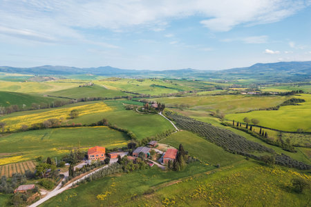 Vast green fields stretch across the landscape, dotted with farms and trees. Rolling hills in the distance create a picturesque view typical of Tuscany during the spring season.の写真素材