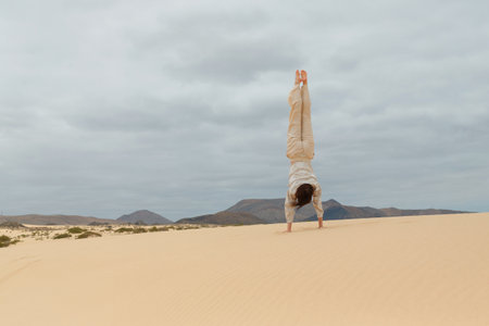 A person dressed in light clothing executes a handstand on sand dunes under an overcast sky in a...