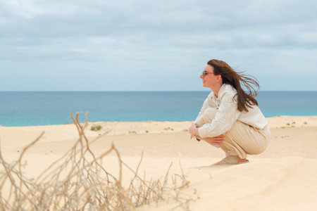 A woman sits thoughtfully on a sandy beach, gazing towards the ocean, with wind in her hair and a...