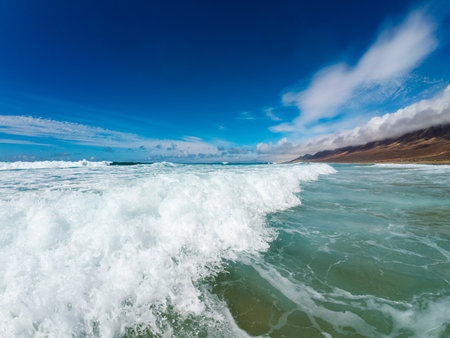 Foamy waves roll towards the shore on a sunny day, creating a tranquil atmosphere at the beach with clear blue skies and distant mountains.の写真素材