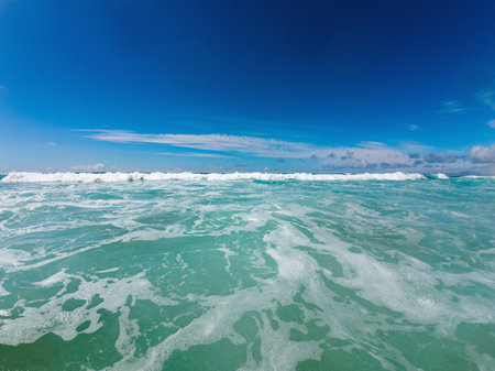 Gentle turquoise waves crash against the shore on a sunny day, with white foam contrasting against the clear blue sky above.の写真素材