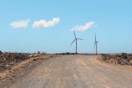 Two large wind turbines rise above a barren landscape near a dirt road under a clear blue sky, showcasing renewable energy in action.の写真素材